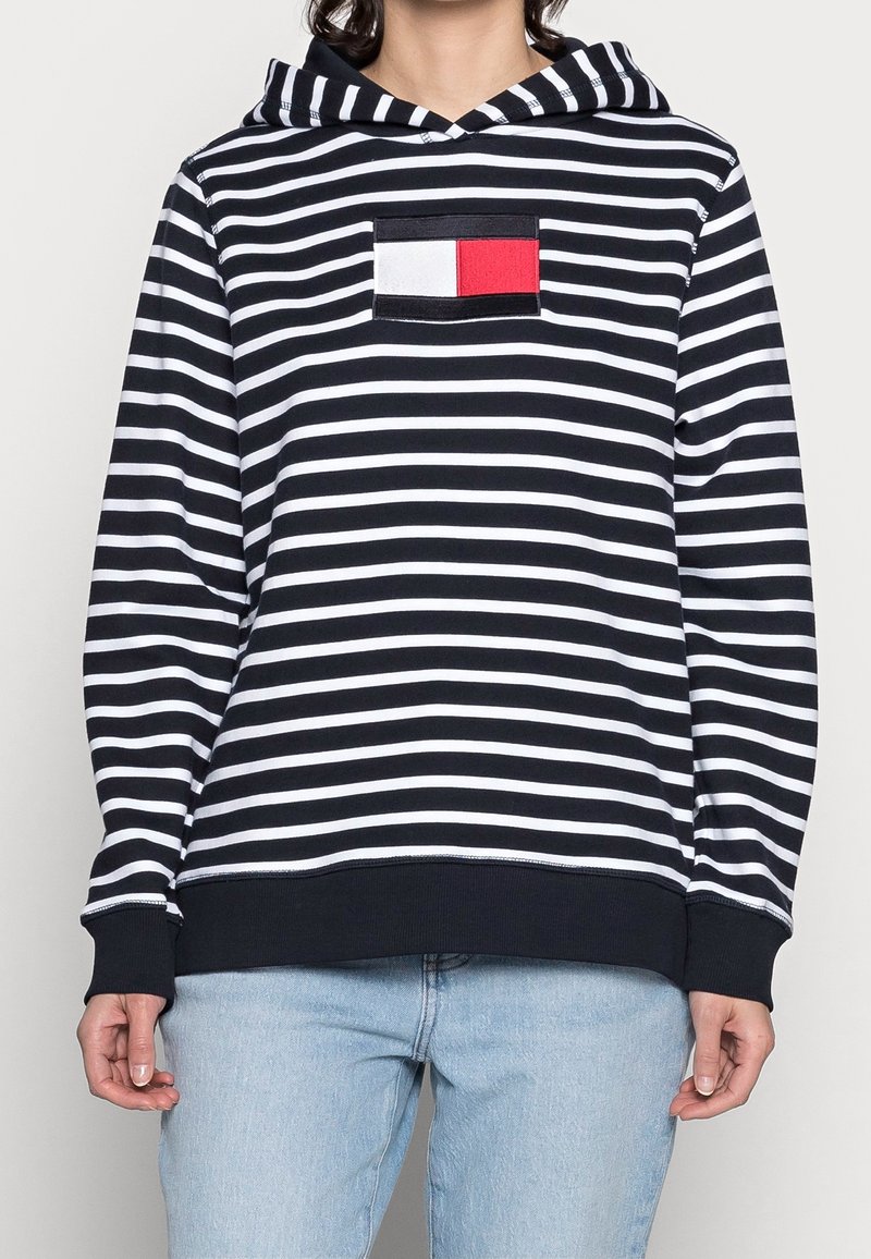 Sudadera con rayas en blanco y negro, con un bolsillo frontal que presenta un parche rectangular rojo y blanco. La tela es suave, de corte casual y con mangas largas.