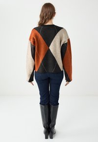 Pull multicolore avec un motif en losange orange, noir et beige ; coupe ample, poignets côtelés et col rond. Jean foncé et bottes jusqu'aux genoux.