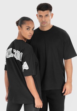 Schwarze, oversized T-Shirts aus weicher Baumwolle, mit einem auffälligen weißen Druck auf dem Rücken. Rundhalsausschnitt und kurze Ärmel, getragen von zwei Models.