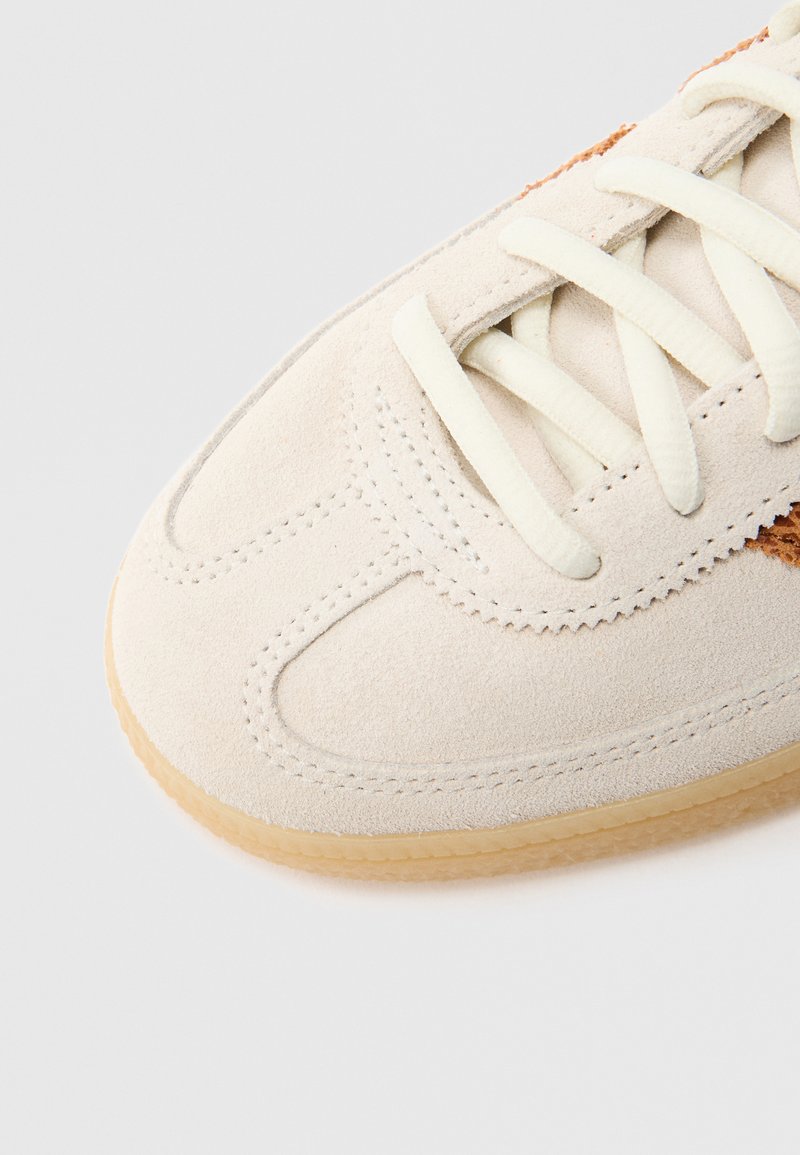 Sneaker in camoscio beige con lacci bianchi, caratterizzata da un design cucito e una suola in gomma naturale con texture. Primo piano che mette in evidenza la punta e la parte superiore.