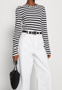 Person in einem gestreiften Schwarz-Weiß-Oberteil mit langen Ärmeln, das in hoch taillierte weiße Hosen mit einem schwarzen Gürtel gesteckt ist, und der eine schwarze Netztasche hält.