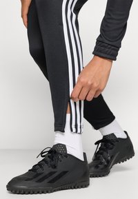 Černé sportovní kalhoty mají bílé trojité pruhy po stranách a na spodní části mají zip. Černé sportovní boty dokončují vzhled.