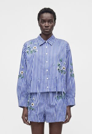 Μοντέλο που φορά μπλε και λευκό πουκάμισο με κάθετες ρίγες και ταιριαστά σορτς με λευκές κεντημένες μαργαρίτες.