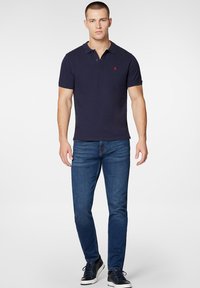 Granatowa koszulka polo z krótkim rękawem, z kołnierzykiem i dwoma guzikami; na piersi wyszywane czerwone logo; zestawiona z niebieskimi dżinsami.