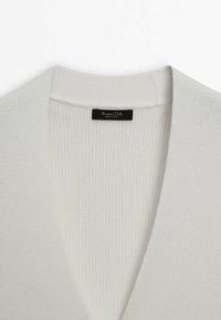 Cardigan bianco lavorato a maglia con profondo scollo a V, texture a coste e sottili punteggiature. Presenta un'etichetta in tessuto nero sul colletto.