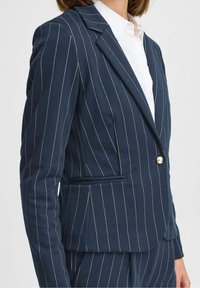 Granatowa marynarka w prążki wykonana z teksturowanej tkaniny, z pojedynczym zapięciem na guzik, klapami z wycięciem oraz boczną kieszenią.