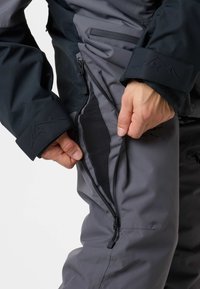 Mani che aprono una cerniera di ventilazione laterale su pantaloni outdoor grigi indossati con una giacca nera che mostra un logo di montagna sui polsini.