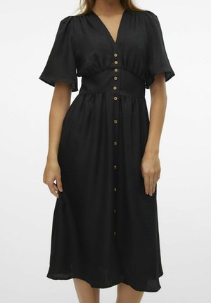 Frau trägt ein schwarzes Midi-Kleid mit V-Ausschnitt, kurzen Ärmeln und einer Reihe Holzknöpfe vorne.