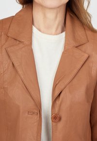 Braune Lederjacke mit Reverskragen, einem Ein-Knopf-Verschluss und einer Brusttasche, getragen über einem weißen Pullover.