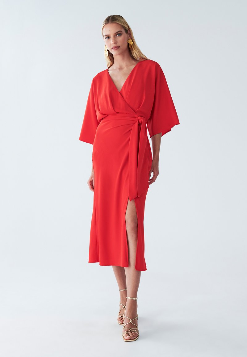 Rotes Wickelkleid mit Kimono-Ärmeln, tiefem V-Ausschnitt, Schlitz vorne und strukturiertem Stoff. Kombiniert mit goldfarbenen Sandalen mit Riemchen.