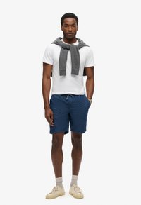 Camiseta blanca, suéter gris atado al cuello, short de baño azul marino con estampado, zapatillas beige y calcetines gris claro.