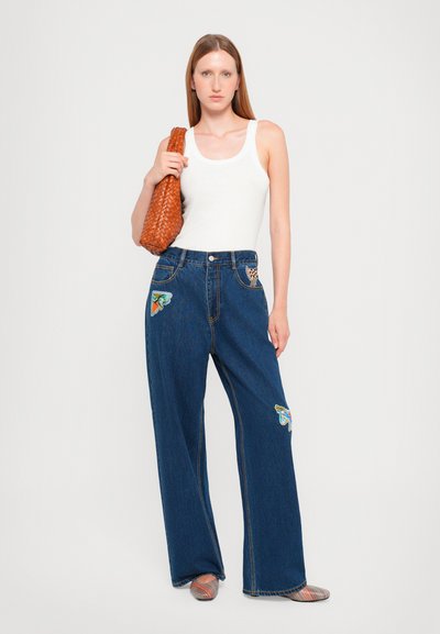 Laiad sinine denim teksad liblikate tikanditega taskutes, koos valge tank topi ja pruuni kootud kottiga.