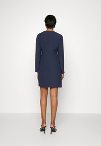 Mörkblå, långärmad klänning med figurnära siluett, texturerat tyg och dragkedja baktill. Stylad med svarta remklackar.