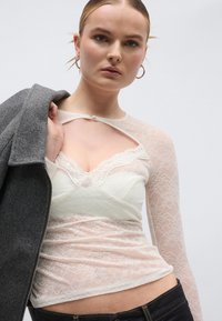 Πλεκτό τοπ με μακριά μανίκια που διαθέτει σχέδιο με άνοιγμα, σε χρώματα ανοιχτό ροζ και ελεφαντόδοντο. Η υφή είναι διάφανη, στρωμένη πάνω από ένα καμησόλ.