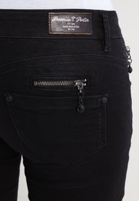 Svarta denimjeans har en läderlogga, en zip-ficka med kedjehandtag och synliga sömdetaljer på baksidan.