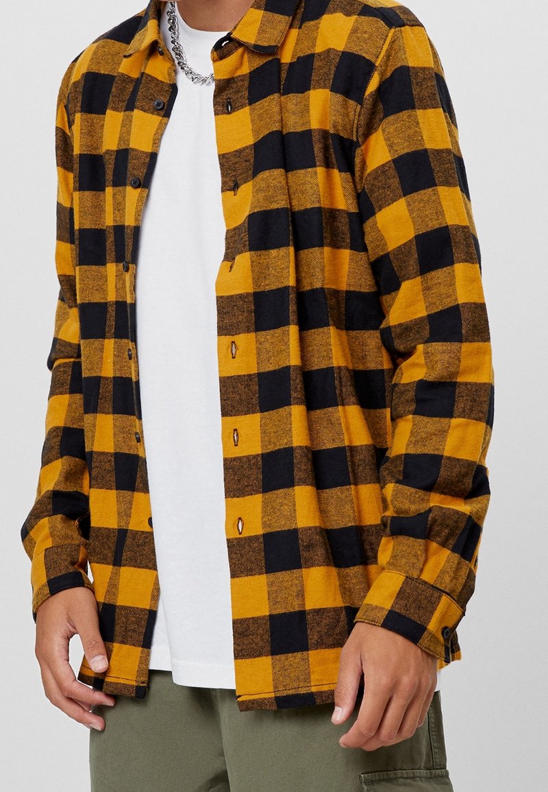 Flanellen overhemd in een geel-zwart ruitpatroon, met knopen, een kraag en lange mouwen, gedragen over een wit T-shirt en groene broek.