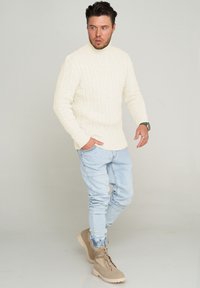 Cremfarbener, kabelgestrickter Rollkragenpullover, hellblaue, schmal geschnittene Jeans und beigefarbene Schnürstiefel. Schlichtes Design mit gerippter Textur und taillierter Silhouette.