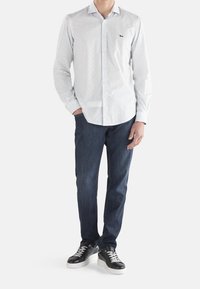 Camicia bianca con piccoli motivi geometrici blu, abbinata a jeans blu scuro e sneakers nere con lacci bianchi.