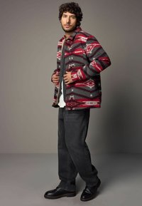 Veste en laine à motif rouge et gris avec un col, portée sur un pull gris et une chemise blanche, assortie à un jean noir et des chaussures.