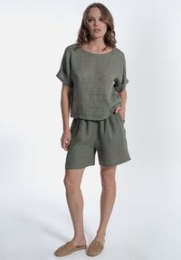 Top in lino verde oliva con maniche arrotolate, abbinato a pantaloni corti coordinati. L'outfit presenta un design rilassato e un tessuto con texture.