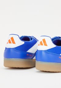 Zapatillas deportivas azules con acentos en blanco y naranja, suelas de goma, y logotipos naranjas en las lengüetas blancas del talón, mostradas desde atrás.
