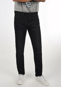 Jeans de mezclilla negra con un ajuste slim, diseño clásico de cinco bolsillos y cierre de botón. Llevados con zapatillas blancas y una camiseta gráfica gris.