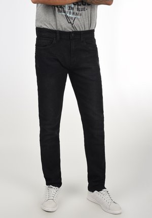 Schwarze Denim-Jeans mit schmaler Passform, klassischem Fünf-Taschen-Design und Knopfverschluss. Getragen mit weißen Sneakers und einem grauen Grafik-T-Shirt.
