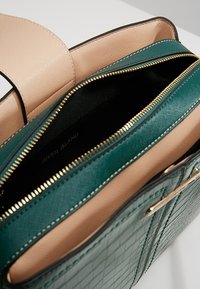 Borsa a tracolla verde con texture, dotata di chiusura a zip, presenta una fodera beige liscia e hardware tondeggianti dorati. Comparti interni visibili.