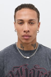Zwart grafisch t-shirt met rode print, zilveren ketting, en meerdere gezichts-tattoos, waaronder een roos en een vlinder, tegen een effen achtergrond.
