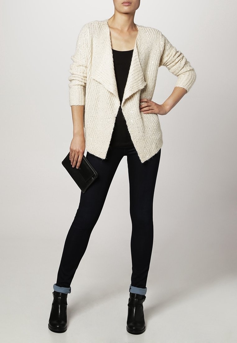 Femme vêtue d'un cardigan en tricot crème par-dessus un top noir, jean skinny foncé avec des revers, bottines noires, tenant une pochette noire.