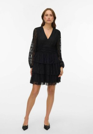 Femme portant une mini-robe à volants en dentelle noire avec manches longues et col en V, associée à des escarpins noirs à bout pointu, debout sur fond blanc.