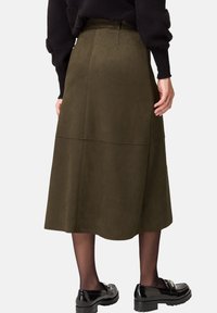 Midi spódnica z zamszu w kolorze oliwkowym, z dopasowaną talią, detalami szwów i rozkloszowanym fasonem, połączona z czarnymi lakierowanymi loafersami i przezroczystymi rajstopami.