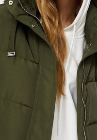 Largo cabello rojo sobre un sudadera blanca debajo de una chaqueta acolchada verde oliva sin abrochar, con botones de presión de metal y cordones.