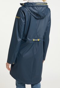 Imperméable bleu marine avec capuche, œillets jaunes et cordon blanc à la taille, texte jaune sur le col et la manche, porté sur un jean.