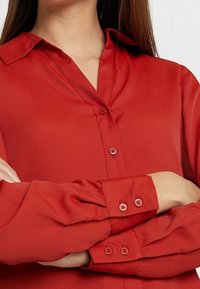 Blouse rouge à boutons avec col, texture douce, manches longues et boutons assortis. Le tissu semble lisse et léger.