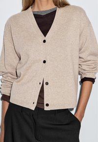 Beige, mjuk stickad cardigan med V-urringning, knappar fram och uppvikta ärmar, över en mörkbrun skjorta, kombinerad med mörka byxor.