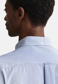 Camicia a righe in cotone bianco e blu, con collo button-down e piega posteriore. Linee pulite e texture liscia.