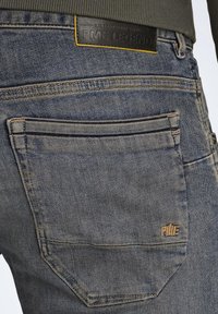 Des jeans en denim bleu foncé présentent une poche latérale avec des coutures discrètes, une étiquette en cuir frappée au logo "PME LEGEND" et une finition texturée.