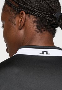 Camisa atlética preta com um colarinho branco. Apresenta um pequeno detalhe de logotipo preto na área da nuca. Textura suave, design ajustado.
