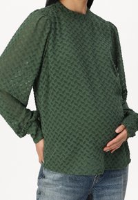 Blouse verte texturée à manches longues, arborant un motif géométrique, un col rond et des poignets légèrement froncés, associée à un jean bleu.