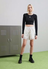 Fekete hosszú ujjú crop top, kis logóval, fehér fodros szegélyű sorttal párosítva. A modellen fekete fűzős csizmák vannak. Szürke záros szekrények a háttérben.