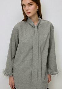 Cămașă gri cu mâneci lungi și guler legat, având un material texturat și accente de pene la manșete. Design simplu, fără model.