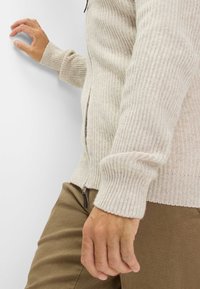 Beige Strickjacke mit Reißverschluss, gerippter Textur und Bündchen. Kombiniert mit braunen Hosen, Hand an einer Wand platziert.