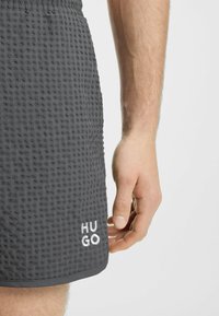 Pantaloni corti grigi in un tessuto testurizzato con motivo a griglia. Il nome del marchio "HUGO" è ricamato in bianco sull'orlo inferiore.