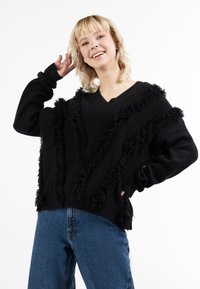 Zwarte oversized trui met V-hals, met franjes aan de voorkant en op de mouwen. Gecombineerd met blauwe spijkerbroek.