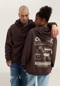 Brown hoodies with kangaroo pockets, featuring white graphic designs of coffee cups and playful text on the back.  Casual, unisex fit.