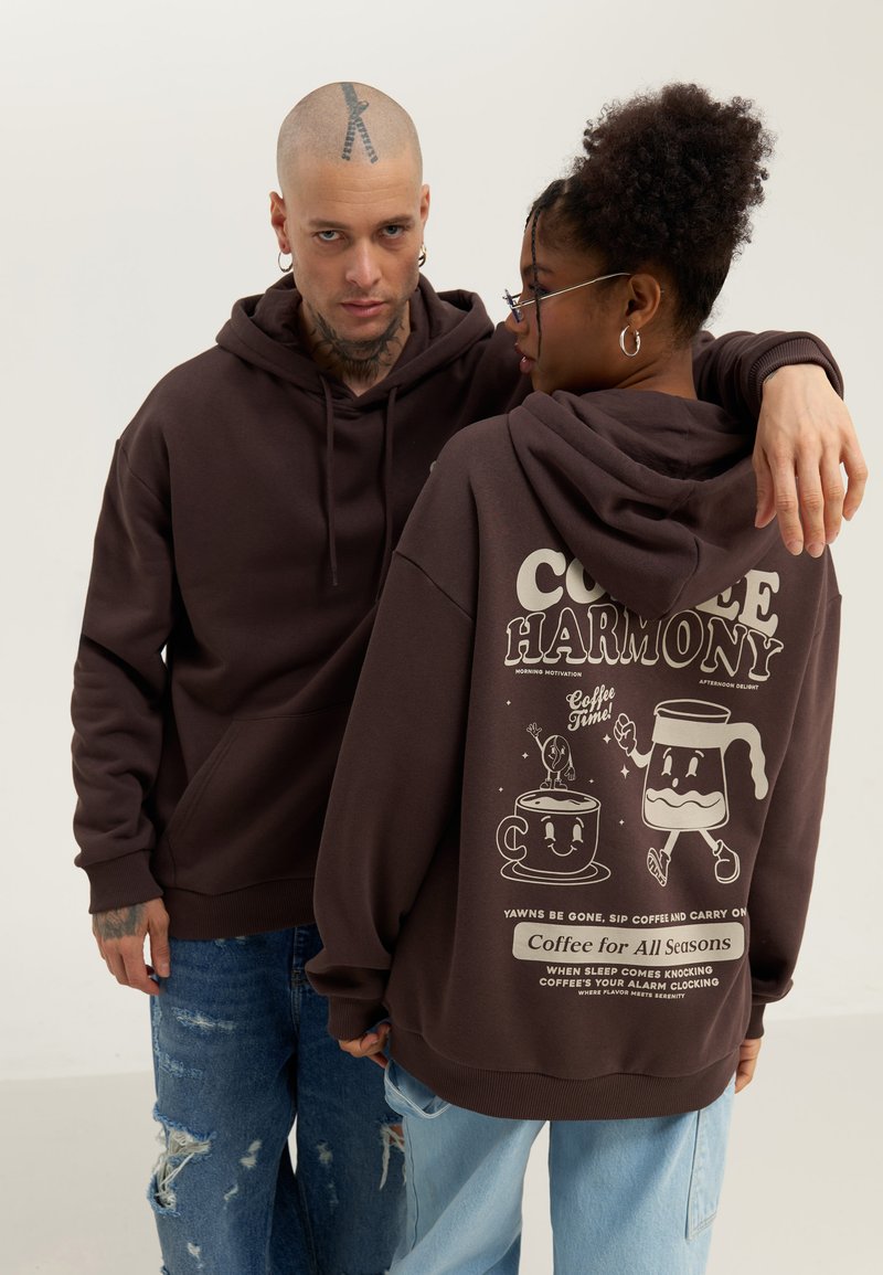 Brown hoodies with kangaroo pockets, featuring white graphic designs of coffee cups and playful text on the back.  Casual, unisex fit.
