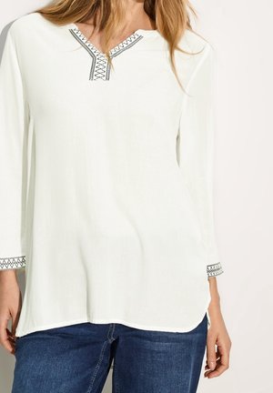 Mujer con túnica blanca de manga larga con bordado geométrico negro en el cuello y los puños, combinada con jeans azules.