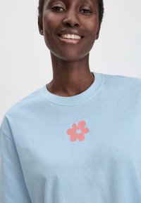 Lichtblauwe katoenen t-shirt met een roze bloemenafbeelding in het midden. Het ontwerp bevat een bloem met zes bloemblaadjes en een vaste kern.