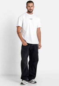 Witte katoenen T-shirt met korte mouwen en groene logotekst op de borst, gecombineerd met zwarte denim jeans en grijze sneakers.